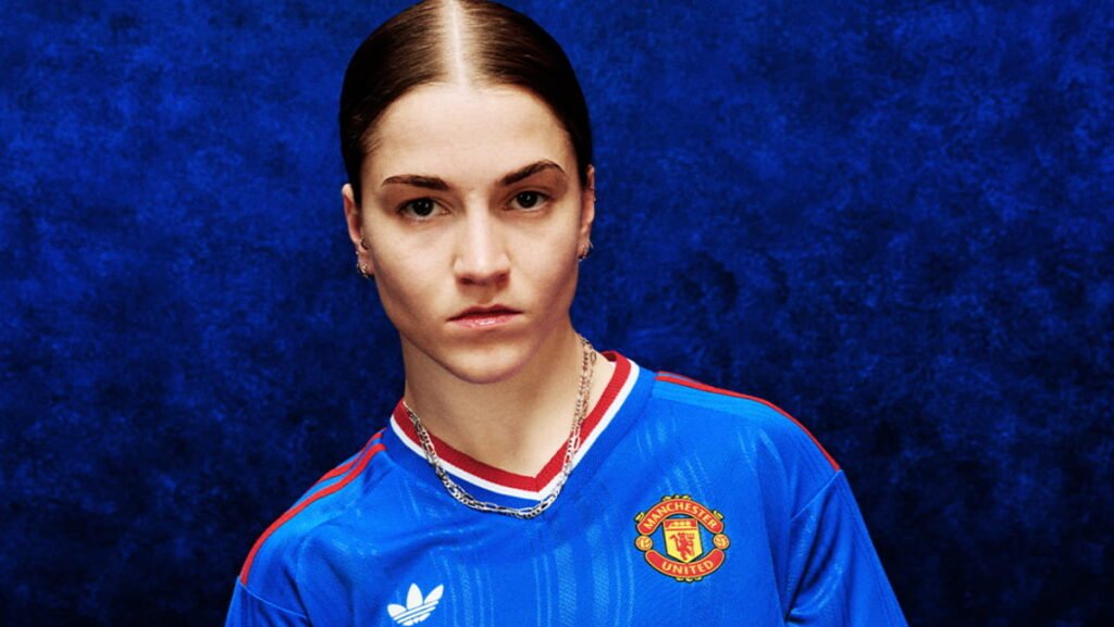 Manchester United Launch Retro Blue Away Kit Ahead of Pre-Season European Tour 2 Footballer Jess Park posing in the Manchester United Football Club blue away shirt with red and white trim and the club badge and Adidas logo on the chest against a blue background