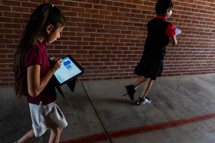 US Schools Reconsider Classroom Screens as States Enact Digital Device Restrictions 2 Two children wearing maroon shirts walk near a red brick wall and look down at iPads.