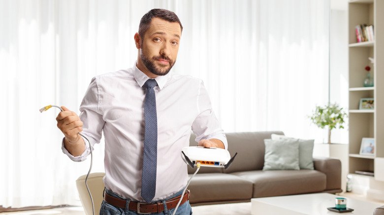 FCC Hardline Ban on Foreign Wi-Fi Routers Stalls Amid Supply Chain Realities 3 A dark haired man in a shirt and tie shrugs as he holds an unplugged Wi-Fi router.