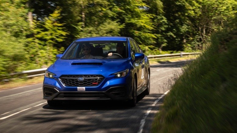 2026 Subaru WRX Base First Drive: Performance Value Purified 8 A head-on shot of a blue WRX driving around a corner on a rural road surrounded by trees and grass