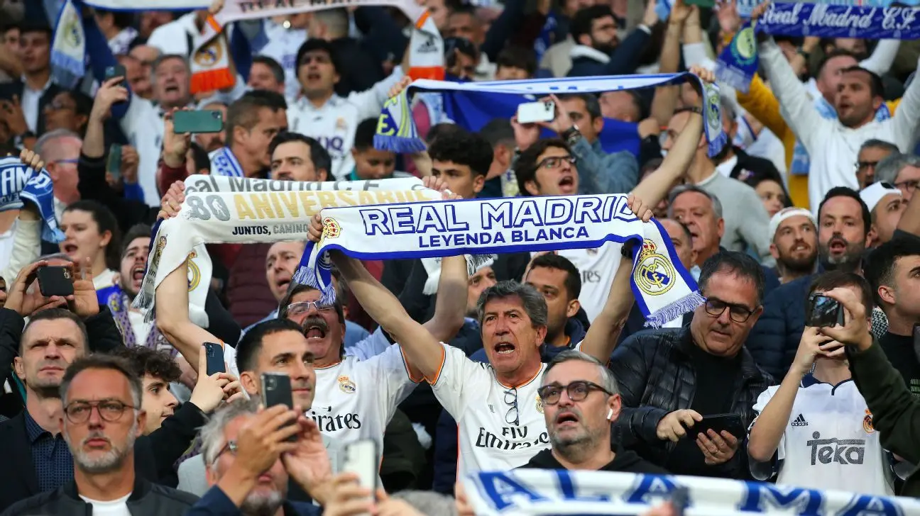 لائحة انضباط جديدة في ريال مدريد تثير غضب الأعضاء وتصادر الانتقادات 2 Real Madrid fans cheering and holding white and blue scarves in the stands of the Santiago Bernabeu stadium