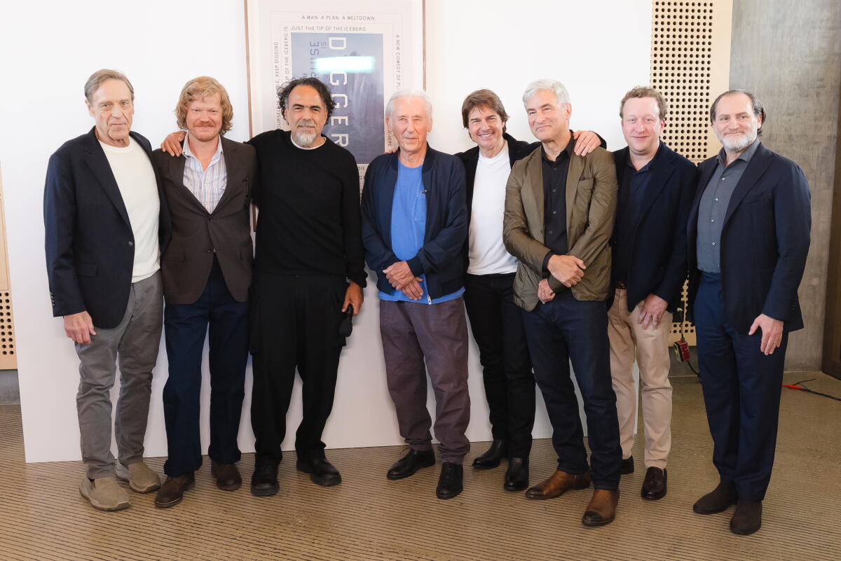 Eight men, in black, brown and blue blazers and suits, pose in front of a framed blue-gray poster on a white wall.