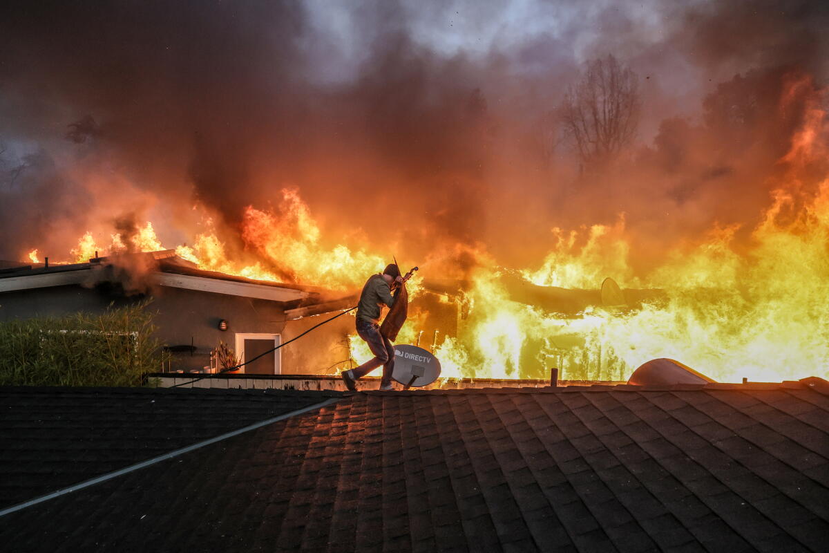 A man on a roof wields a garden hose with flames in the background.