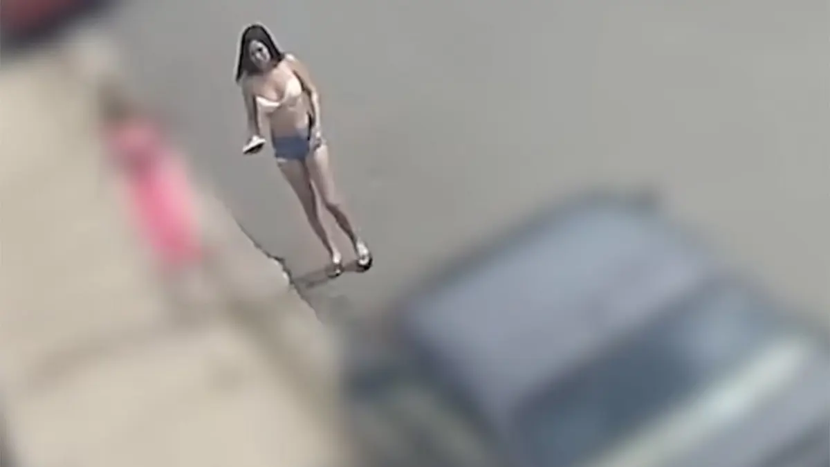 A woman in a light-colored top, denim shorts and sandals faces the camera on a paved street, holding an object in one hand.