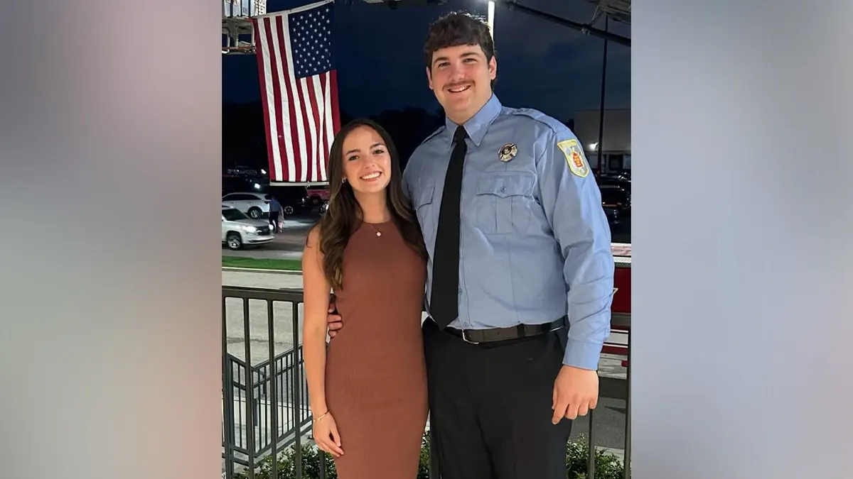 Firefighter Shane Hirstius, wearing a light blue uniform shirt and black tie, stands with his fiancée, Leah Fonte, in a brown dress beneath a large American flag.