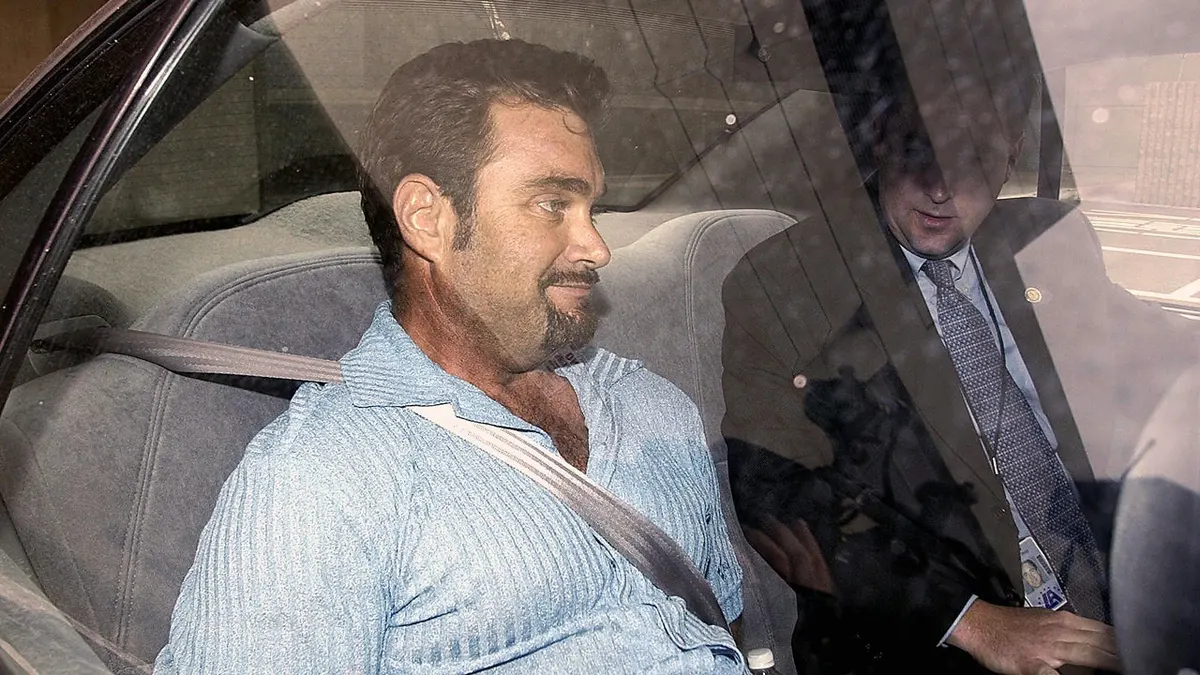 Andrew Luster, wearing a light blue shirt and a seat belt, sits in the back seat of a vehicle beside a suited law enforcement official.