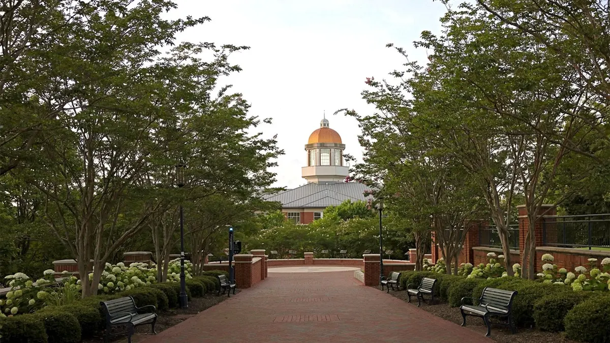 A tree-lined brick walkway with benches and landscaping leads to a domed building on the University of North Carolina at Charlotte campus.