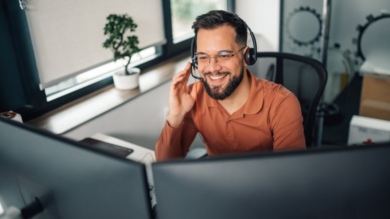 A man wearing a headset and smiling at his desk with two monitors visible in the foreground.
