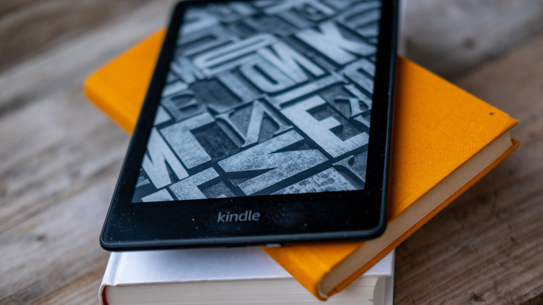 A Kindle laying on top of a couple of books, on a wooden table.